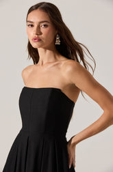 A woman is wearing a strapless black dress with a fitted bodice and a pleated skirt. She has long brown hair and is accessorized with large, dangling pearl earrings.